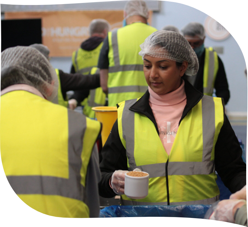 An Asian staff member works with her colleague at a rice packing event. They are both wearing yellow vests and hair nets.