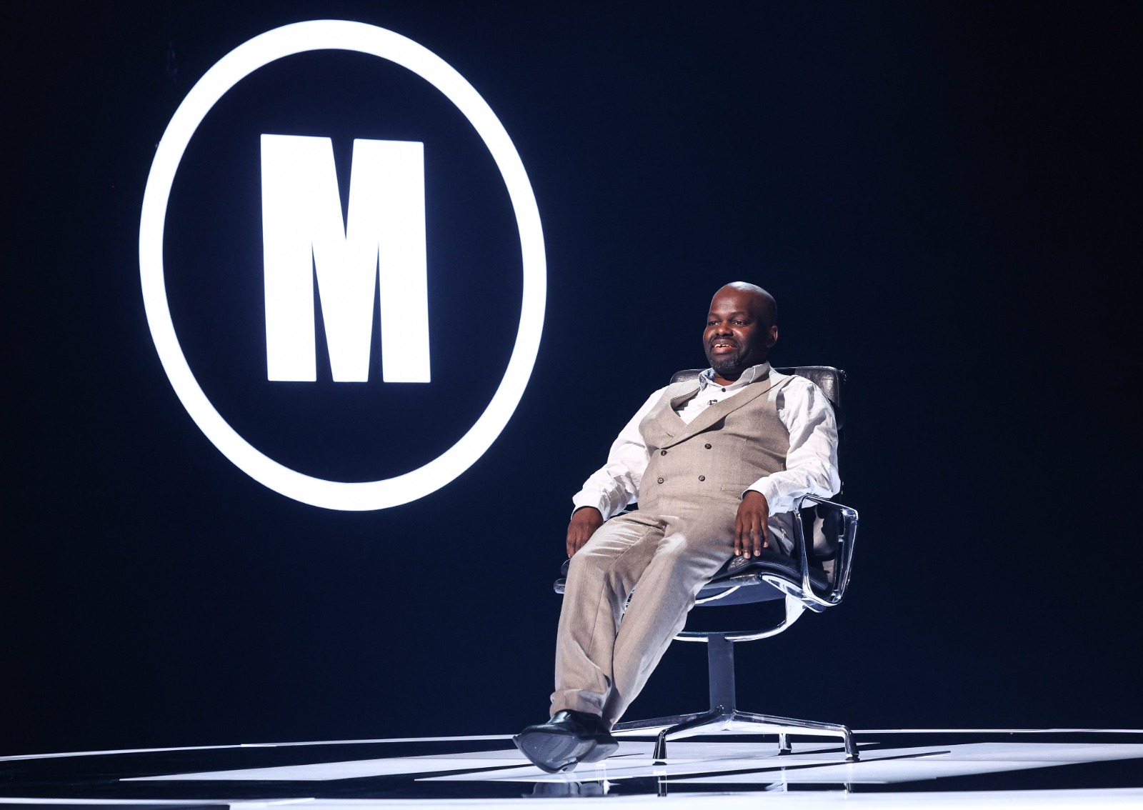 Black comedian Daliso Chaponda sitting on a black and silver chair. He is on the Mastermind set. Behind him is the large 'M' logo of the show. He is smiling.