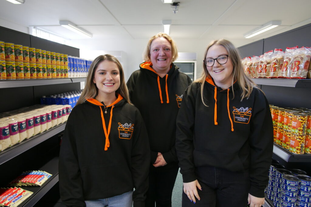 Three women stand together in the centre of a supermarket aisle. They are all looking at the camera with broad smiles on their faces. They are wearing black and orange hoodies branded with The Community Pantry logo.