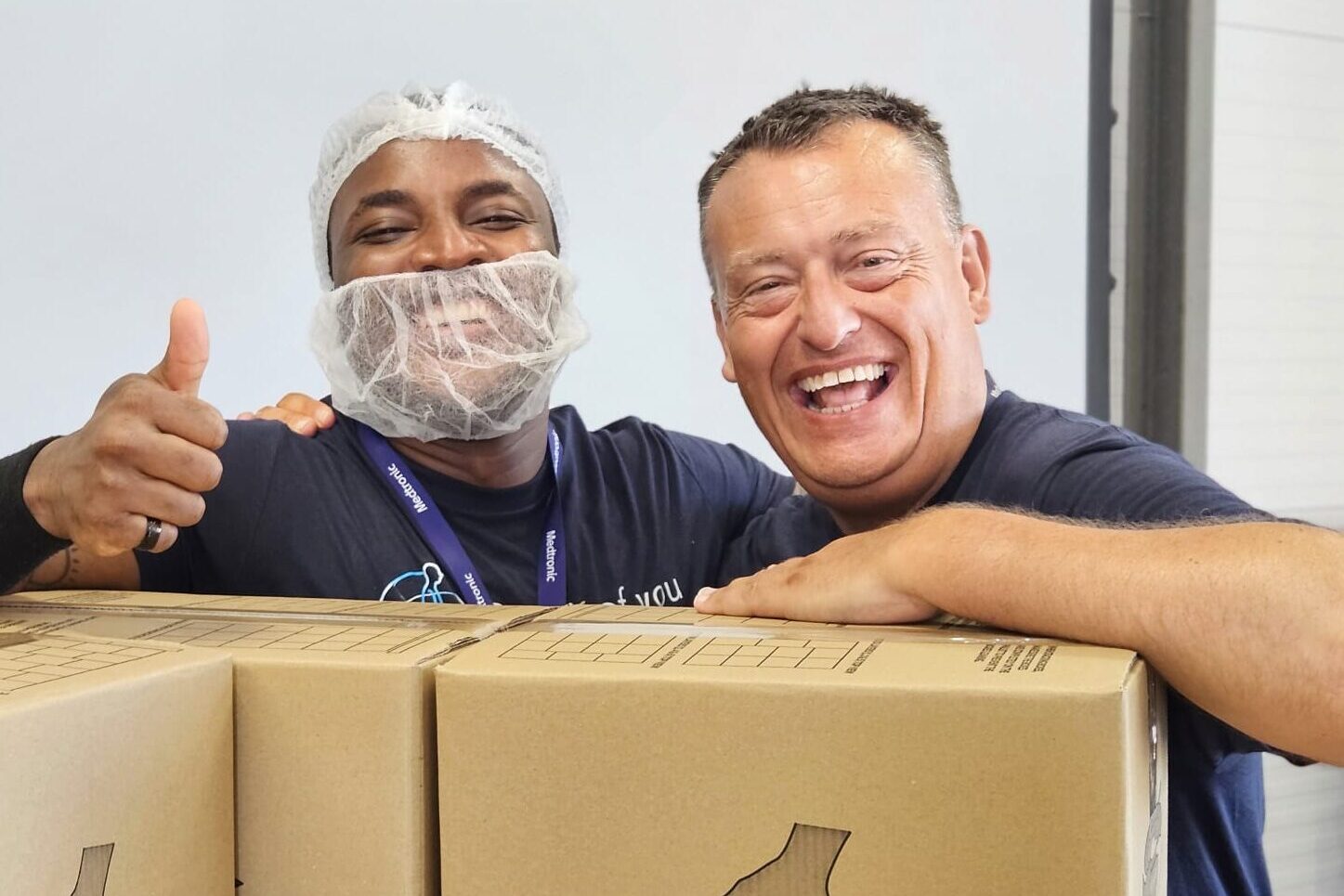 Two men grin at the camera. One man is wearing a catering hair net and giving a thumbs up. They are leaning on boxes packed with rice meals.