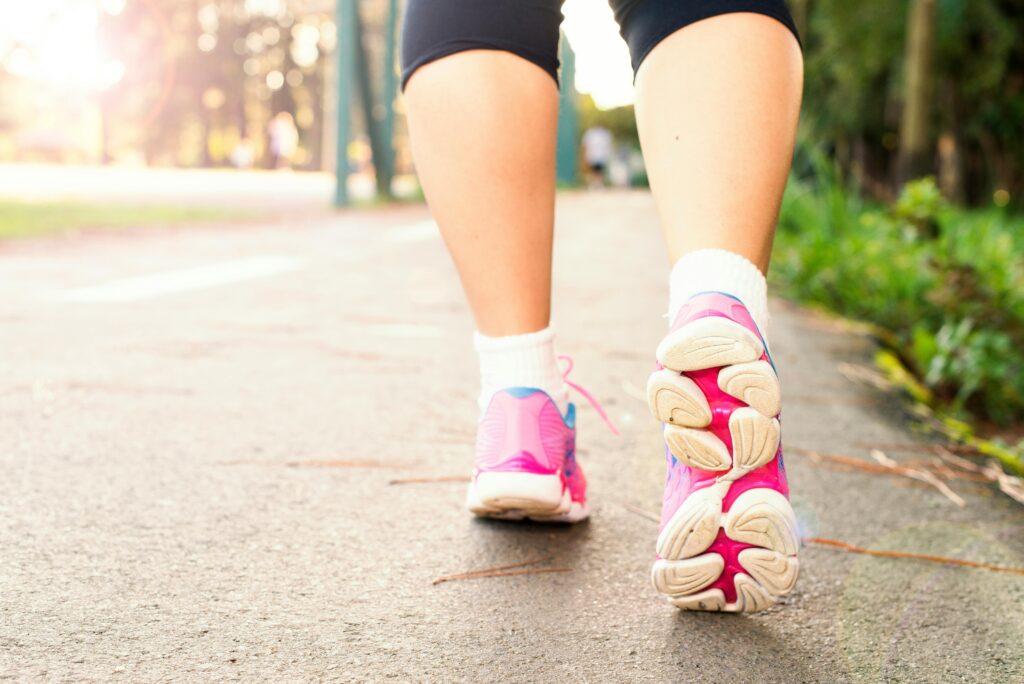 A woman's pink and white running trainers. She is wearing fitness leggings and walking along a path lined with grass.